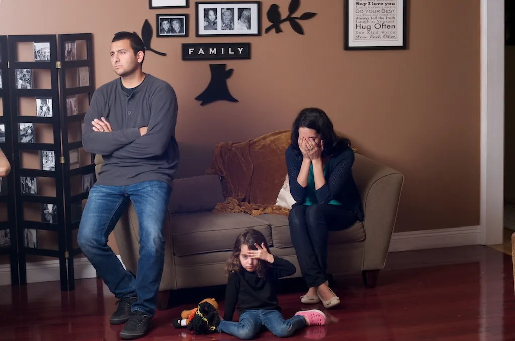 Husband and wife sit on the couch, their daughter upset on the floor.